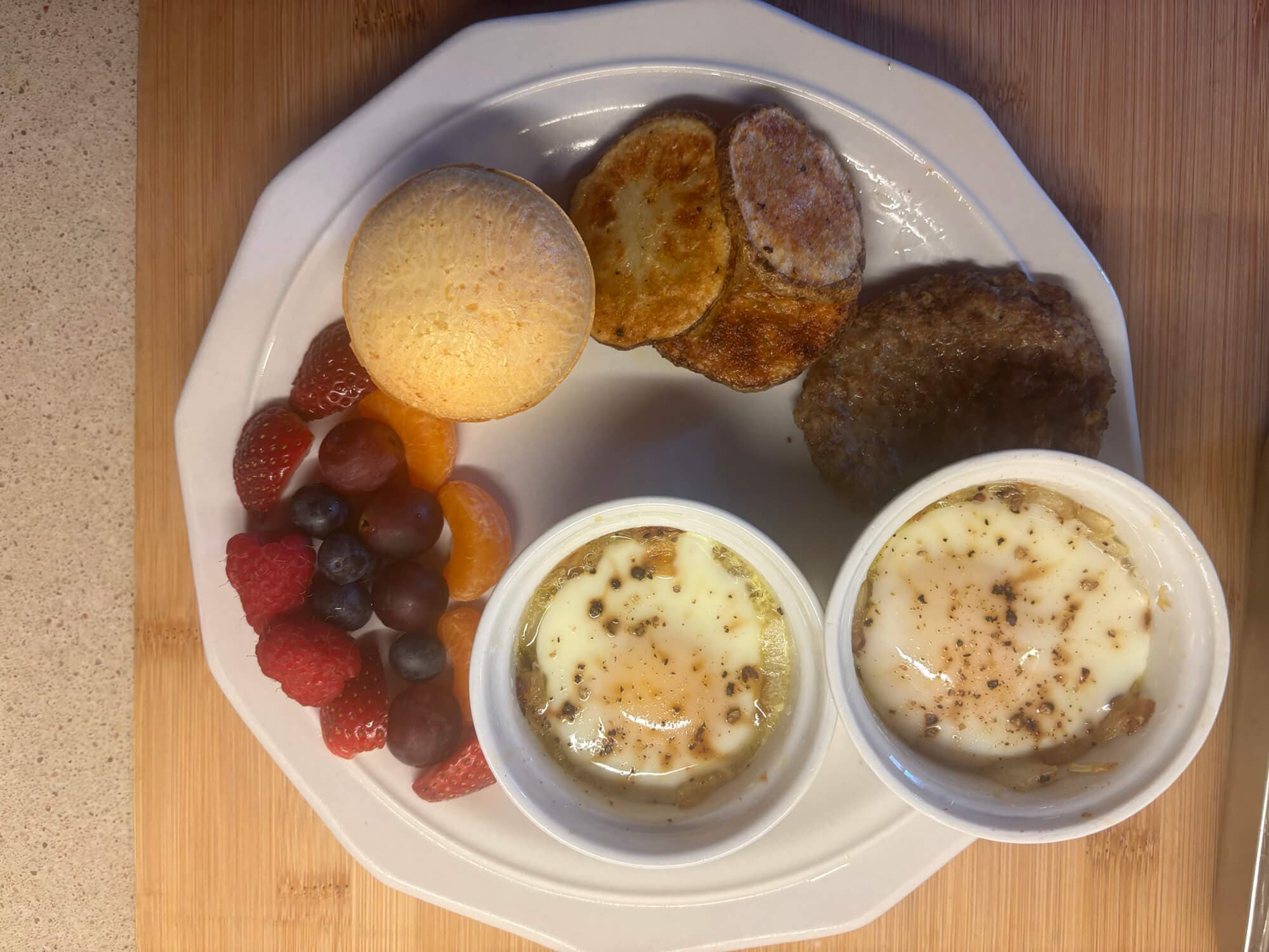 A plate featuring two baked egg cups, a muffin, sausage patty, toasted bread, and a variety of fresh fruit.