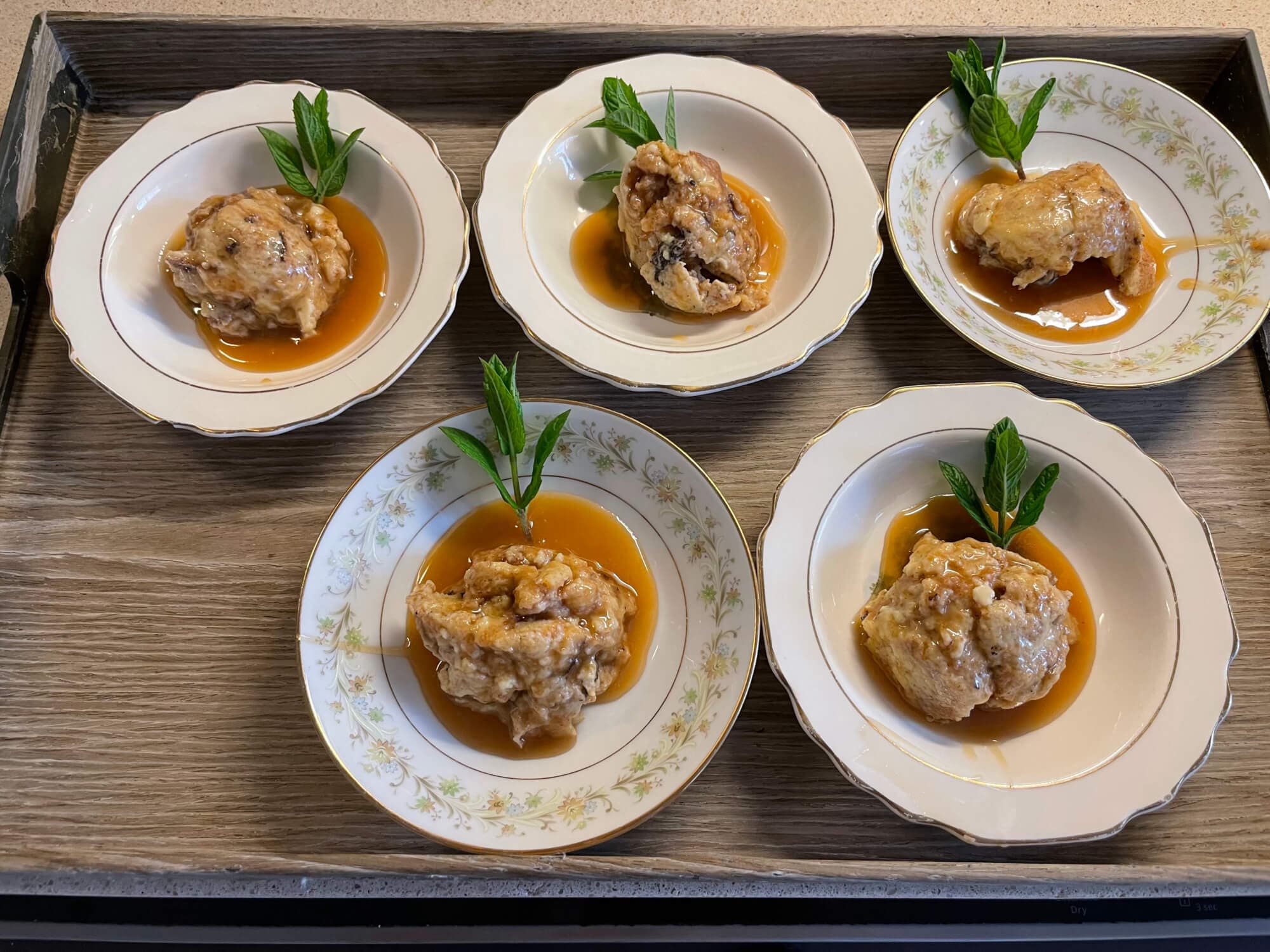 Five dishes of dessert served on plates, each topped with mint leaves and drizzled with a sweet sauce, arranged on a wooden tray.