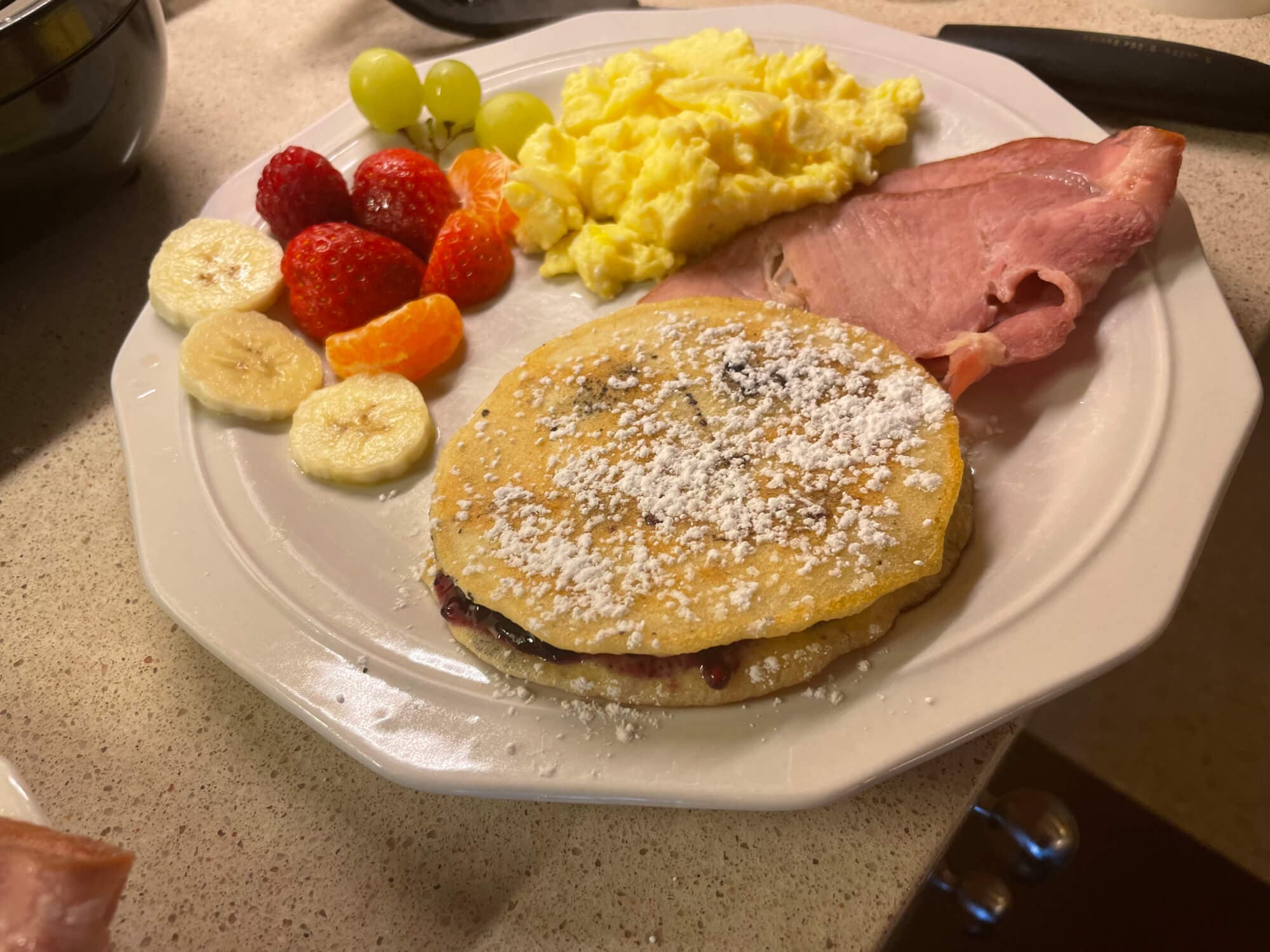 A plate of breakfast featuring a pancake topped with powdered sugar, scrambled eggs, ham, and a variety of fruits.