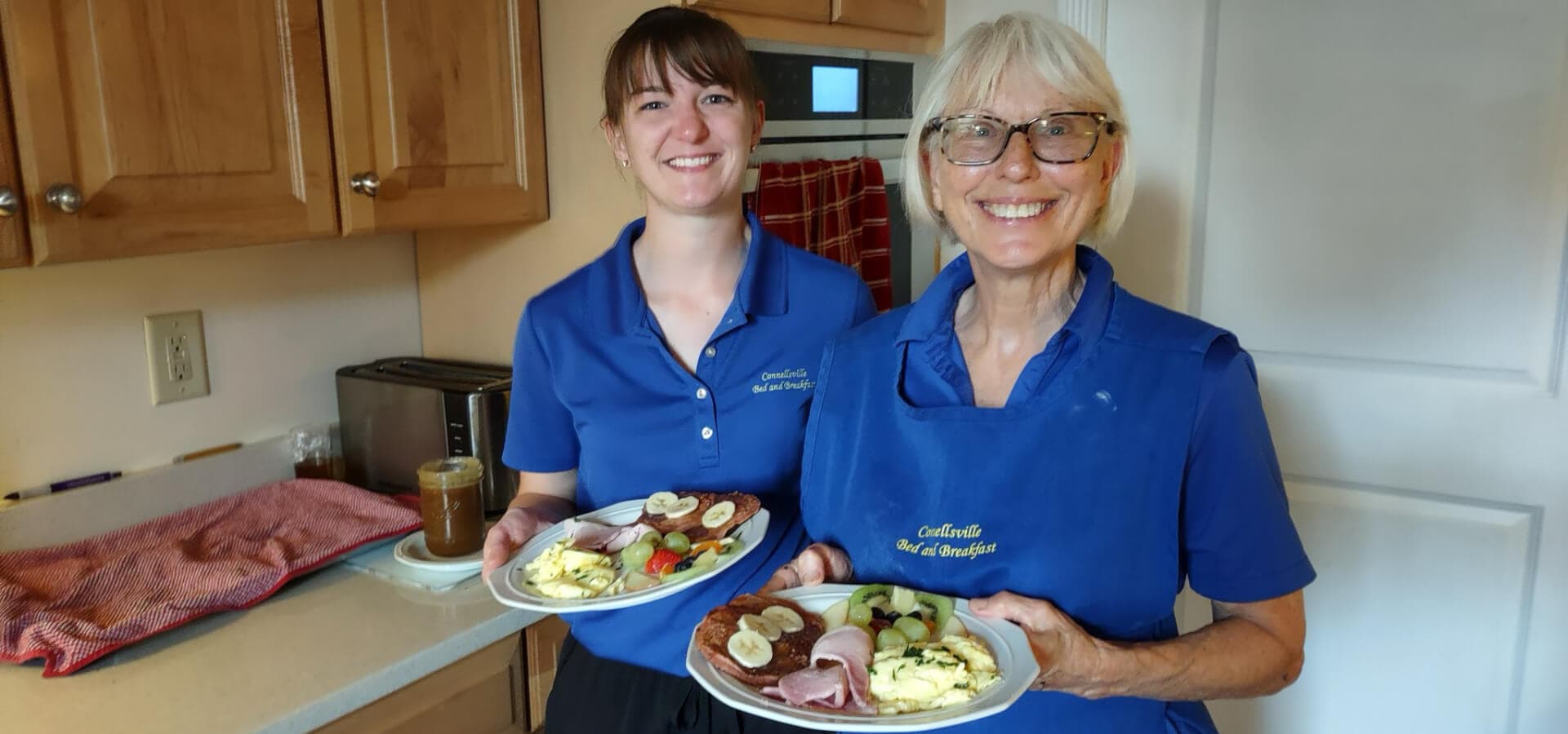 Two women in matching blue shirts smile while holding plates of breakfast food in a kitchen.