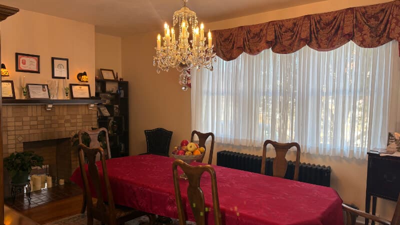 A warmly lit dining room featuring a red tablecloth, wooden chairs, a chandelier, and a window with sheer curtains.