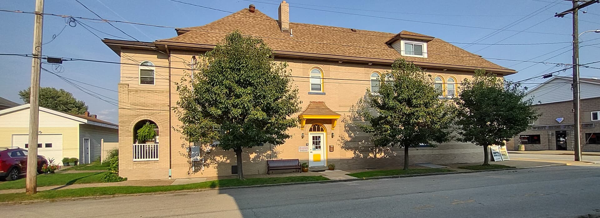 A two-story brick building with a yellow door, flanked by trees, sits on a quiet street.