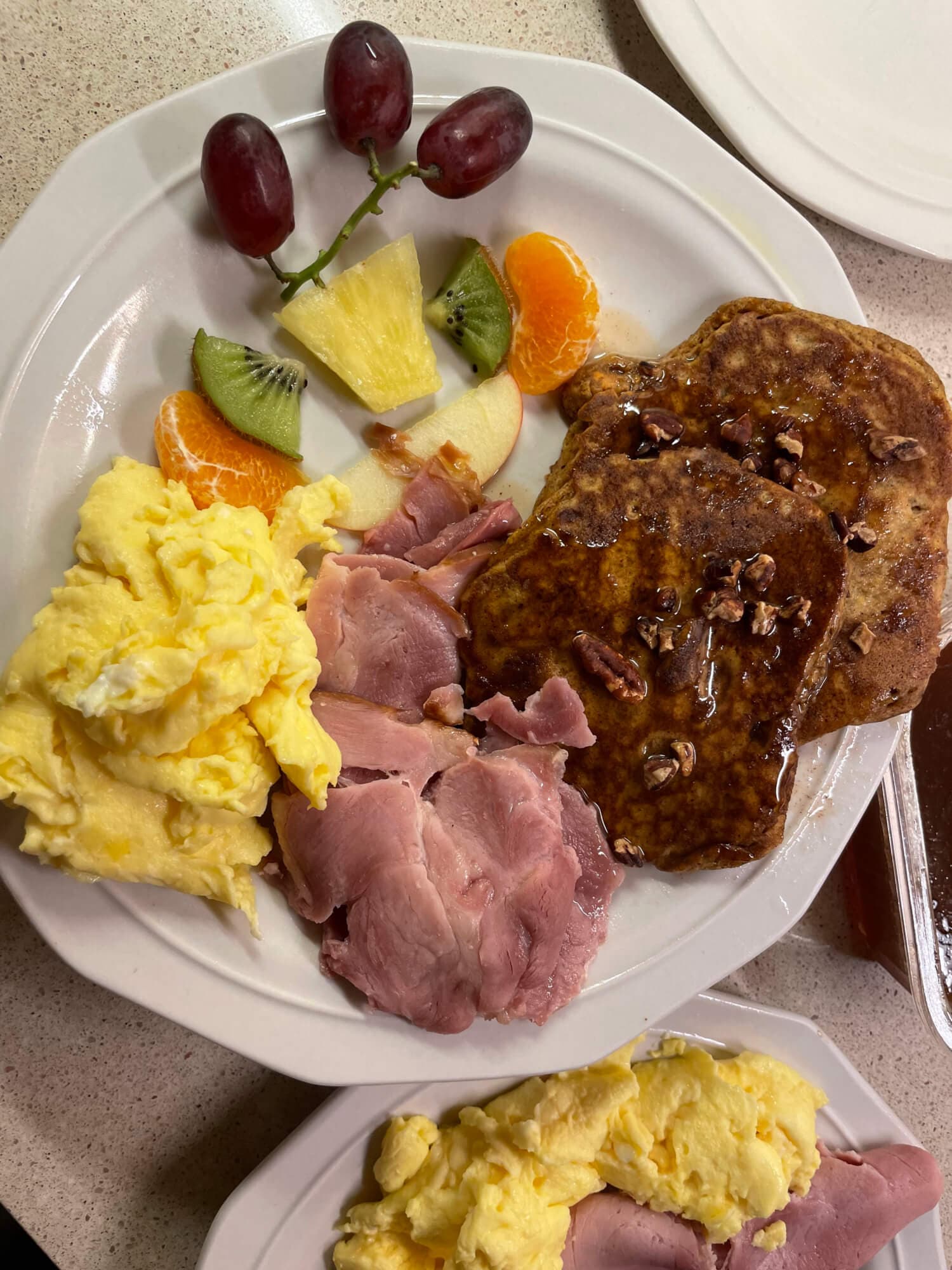 A breakfast plate with scrambled eggs, sliced ham, pecan pancakes topped with syrup, and assorted fruits.