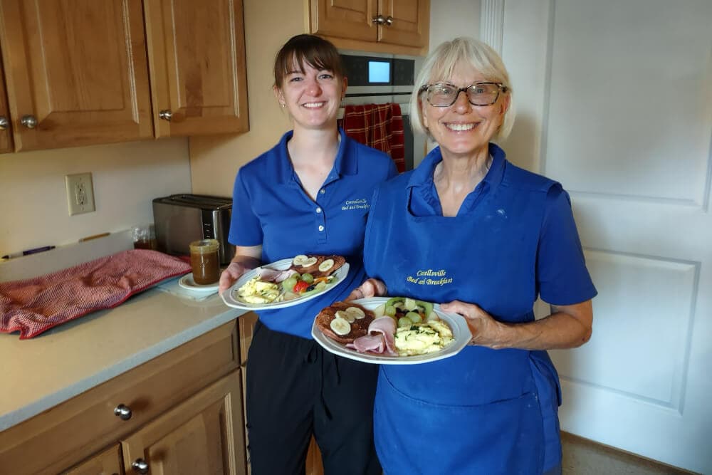 Two smiling women in blue shirts hold plates of food in a kitchen.