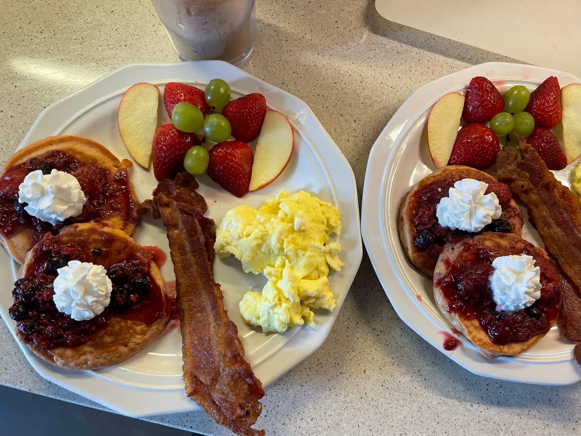 Two plates of breakfast featuring pancakes with fruit topping and whipped cream, scrambled eggs, bacon, and fresh fruit.