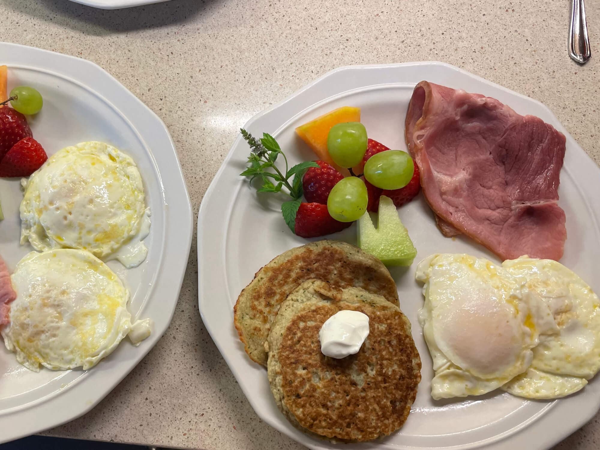 Two plates of breakfast featuring eggs, ham, pancake-like patties, and assorted fresh fruits.